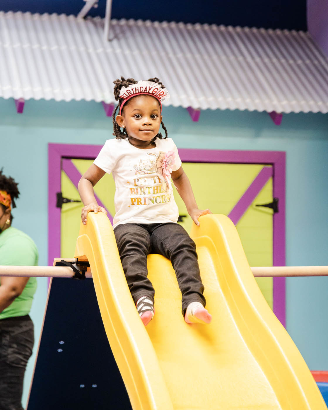 A girl with a birthday girl crown sliding down a yellow slide at Romp n' Roll's kids birthday parties.